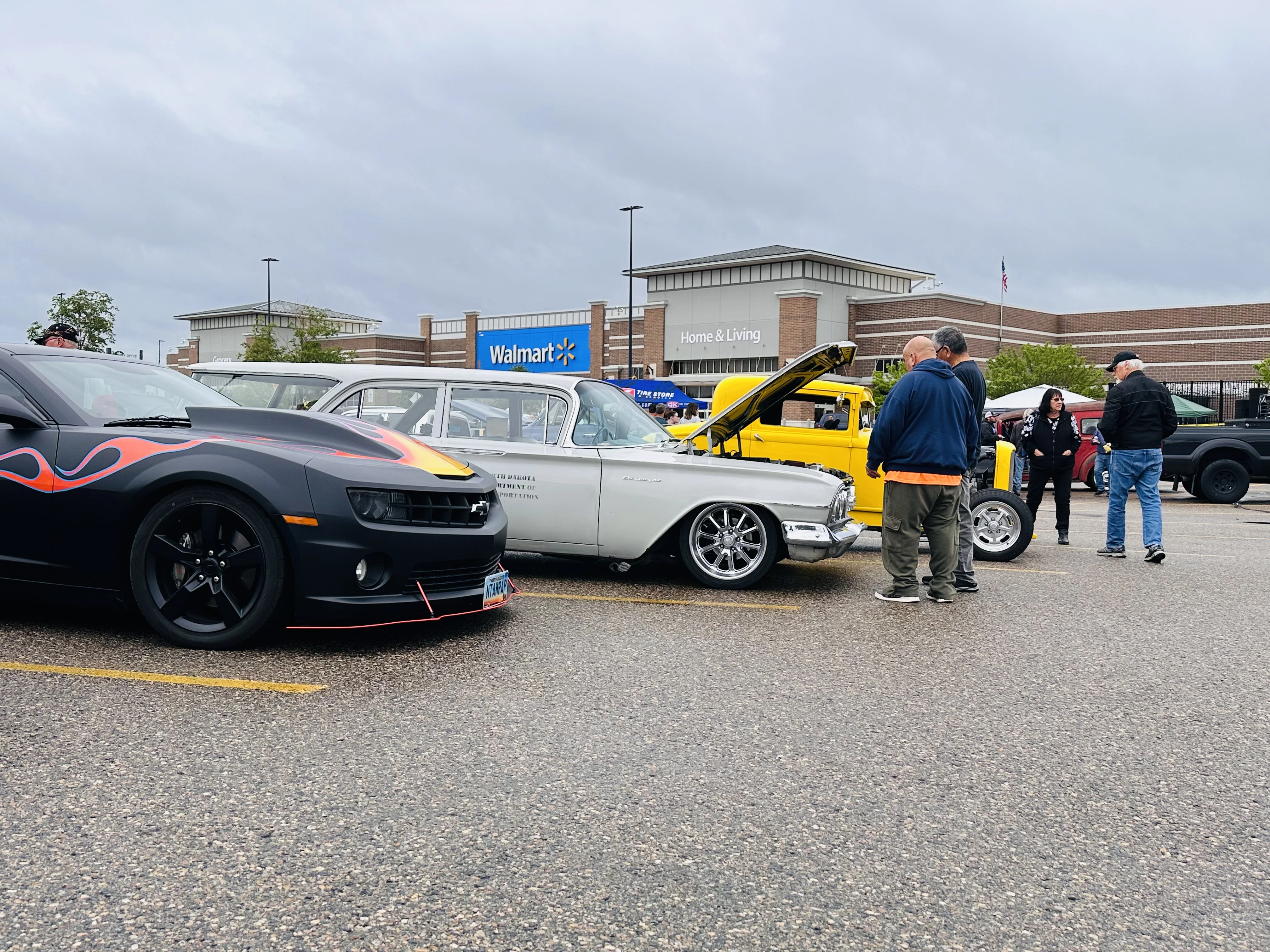 Flame-painted Camaro and silver classic car parked in front of Walmart with a yellow custom truck