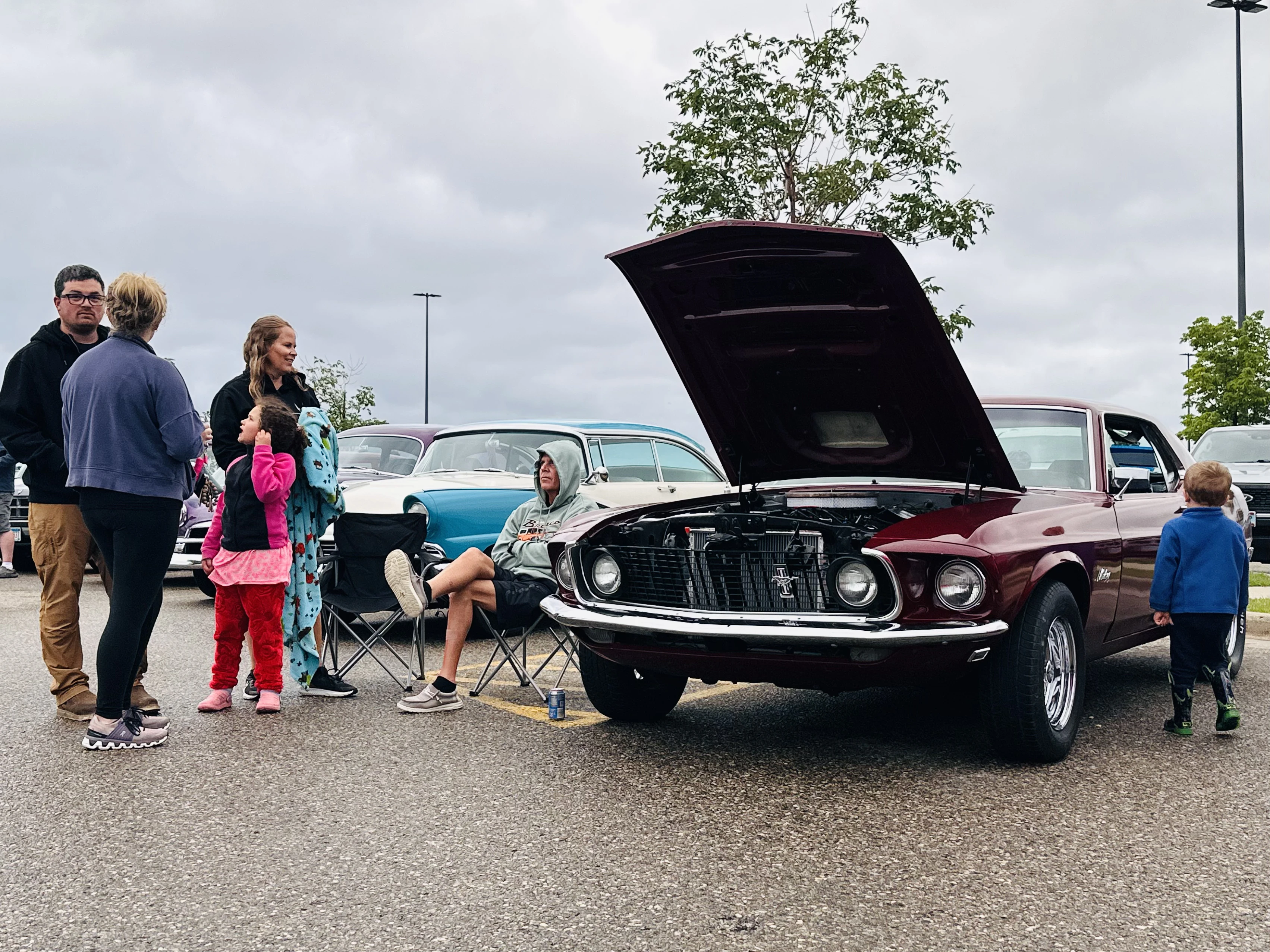 Family with young children gathered around a classic Mustang with its hood up