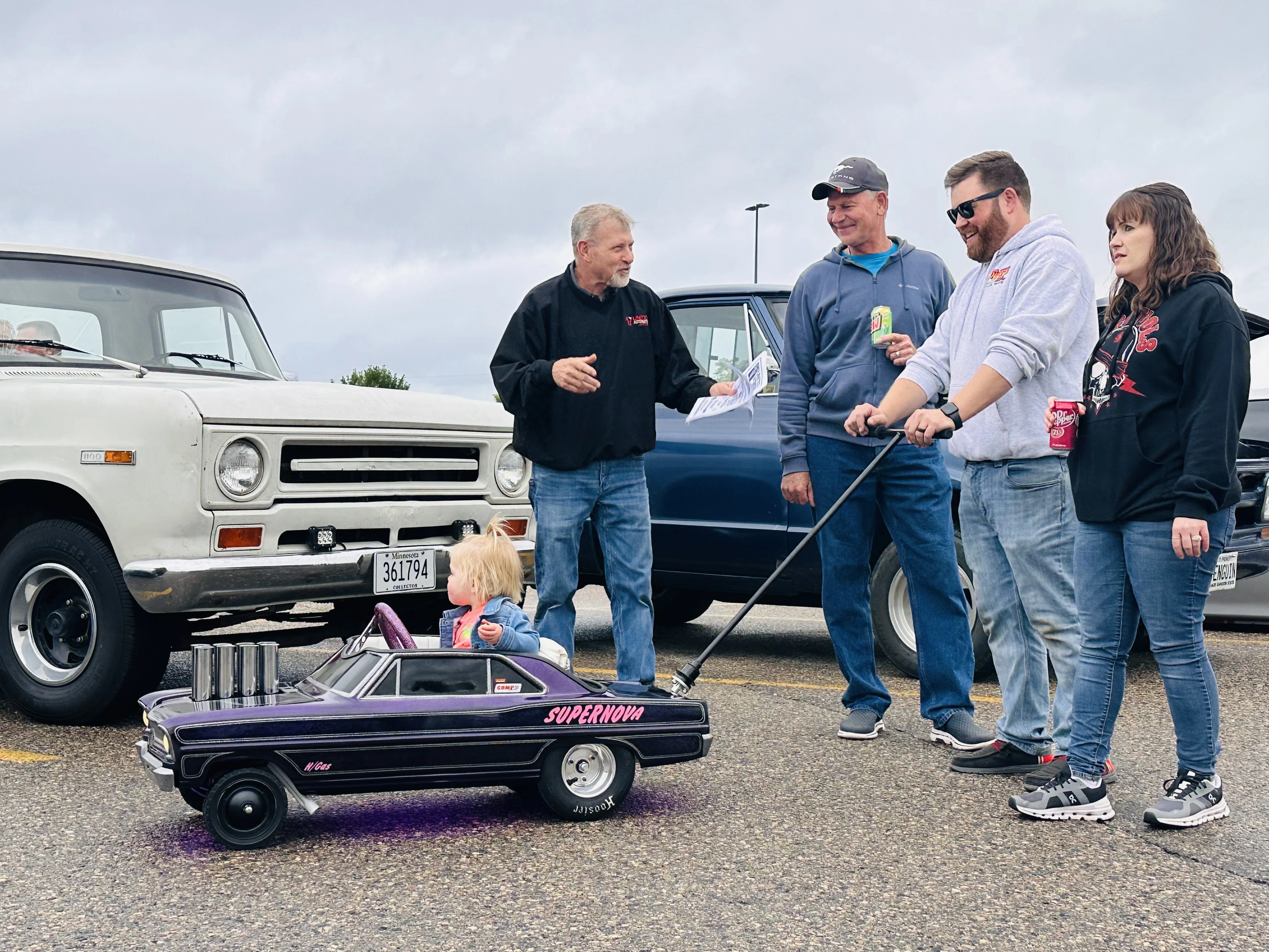 Toddler riding a purple Supernova pedal car while adults chat among classic trucks